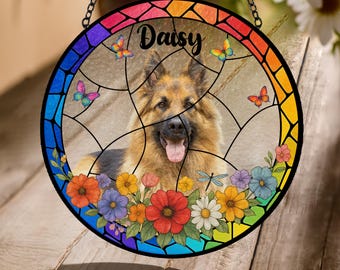 May include: A stained glass suncatcher with a rainbow border and a German Shepherd dog in the center. The dog is surrounded by flowers and butterflies. The text "Daisy" is written at the top of the suncatcher.