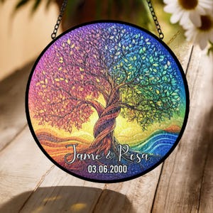May include: A round, stained glass window featuring a colorful tree design. The tree's leaves transition through a rainbow of colors. The names "Jame & Rosa" and the date "03.06.2000" are written on the bottom. The window is framed in black and hangs from a chain.
