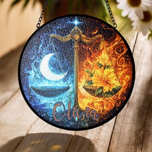 May include: Round stained glass art piece featuring a balance scale design. One side depicts a crescent moon in blue tones, the other a flower in fiery orange hues. The name "Olivia" is written in cursive.