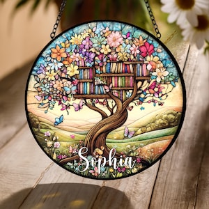 May include: Round stained glass window art featuring a tree with bookshelves for branches, colorful flowers, and butterflies. The name "Sophia" is written in cursive script. The artwork is framed in black and hangs from a chain.