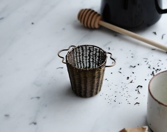 Puede incluir: Un pequeño infusor de té de color latón con una cesta de malla tejida y dos asas. El infusor está sobre una superficie de mármol blanco.