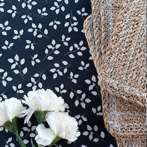 May include: A still life arrangement with three white carnations and green stems, set against a dark blue fabric patterned with white leaves. Woven placemats are visible on the right, adding texture and contrast.