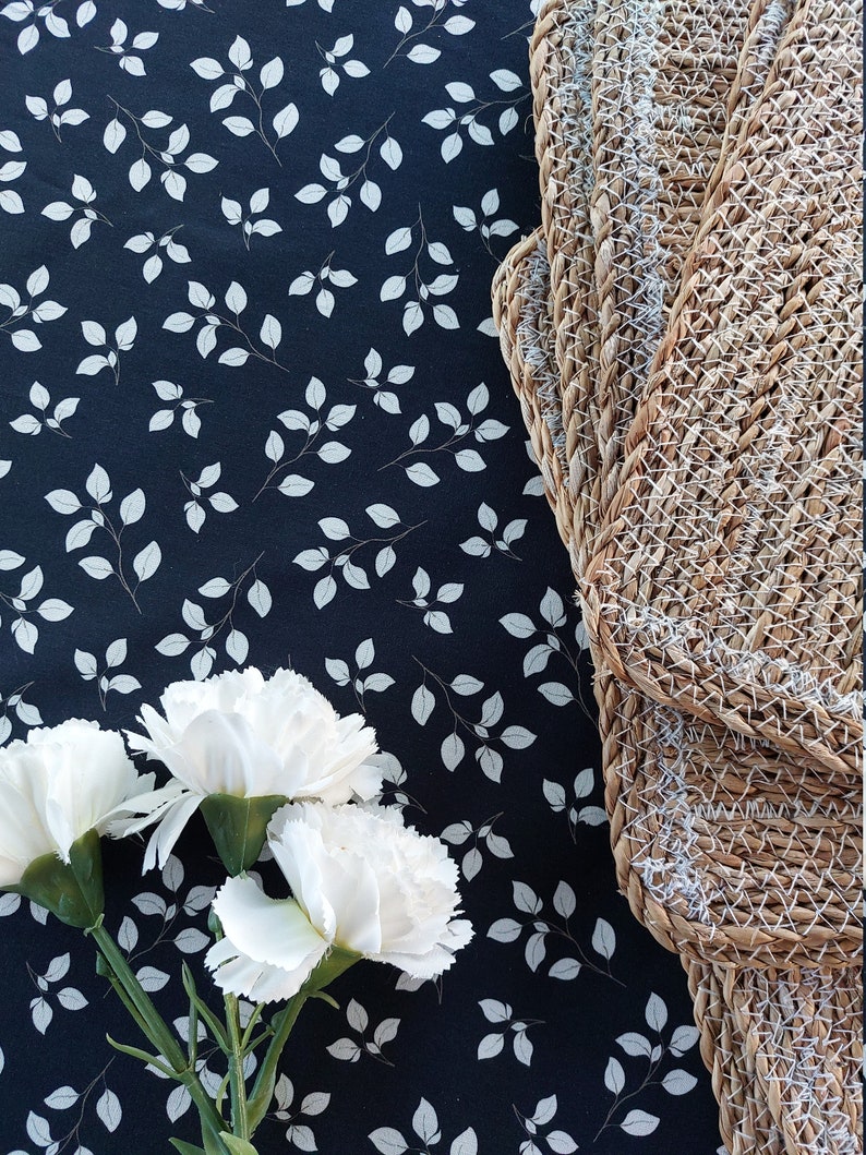 May include: A still life arrangement with three white carnations and green stems, set against a dark blue fabric patterned with white leaves. Woven placemats are visible on the right, adding texture and contrast.