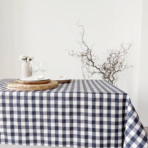 May include: A navy blue and white checkered tablecloth covers a table set for dining. The table is set with white plates, wine glasses, and a vase of white flowers. A decorative branch is in the background.