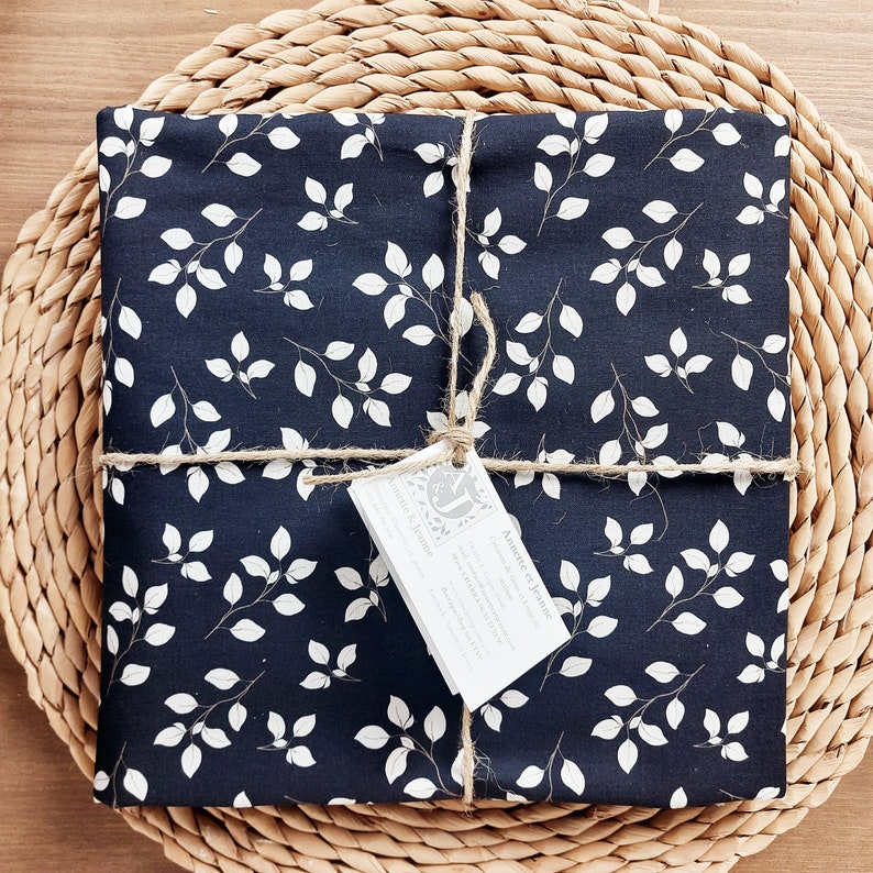 May include: Navy blue fabric with a white leaf design, neatly folded and tied with twine. A small tag is attached. The fabric is presented on a woven, circular placemat.