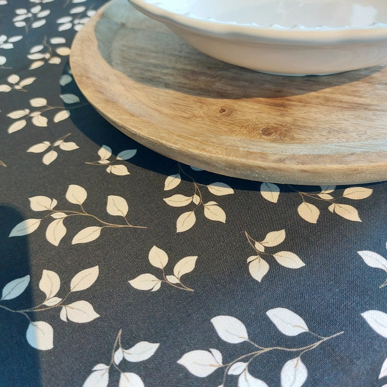 May include: A close-up of a dark grey tablecloth featuring a repeating pattern of white leaves and tan stems. A wooden charger and a white bowl with a scalloped edge are on top of the tablecloth.