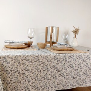 May include: A white tablecloth with a floral pattern, two place settings with white plates, wooden placemats, and wooden bowls. A glass candle holder and a glass of water are on the table. A vase with dried flowers is on the table.