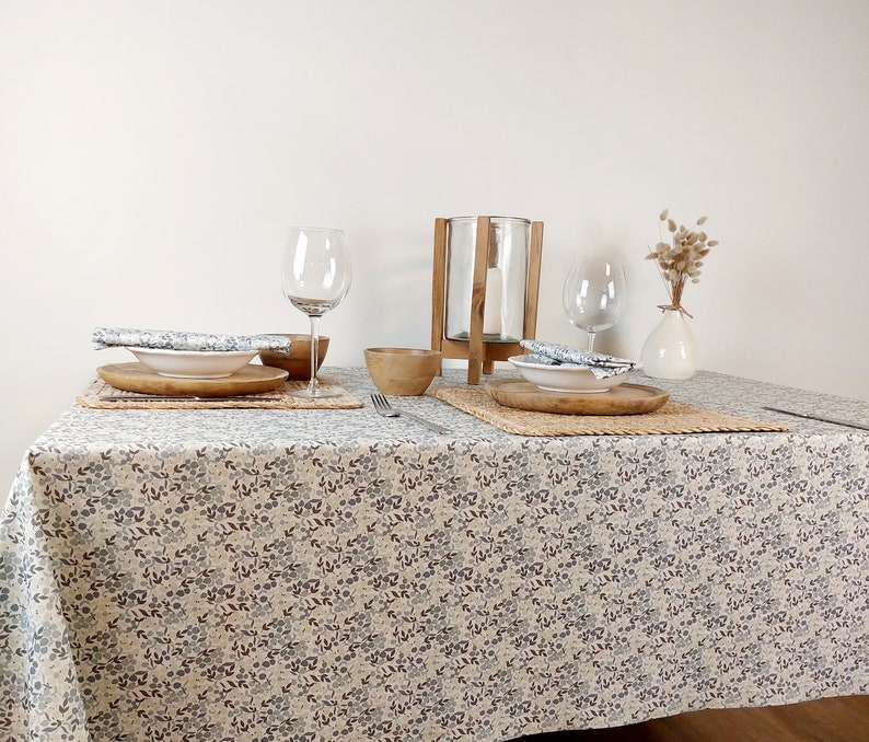 May include: A white tablecloth with a floral pattern, two place settings with white plates, wooden placemats, and wooden bowls. A glass candle holder and a glass of water are on the table. A vase with dried flowers is on the table.