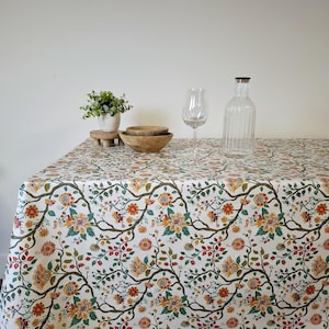 Puede incluir: Un mantel blanco con un estampado floral con flores naranjas, amarillas y rojas, hojas verdes y ramas marrones. Una botella de vidrio transparente, una copa de vino y cuencos de madera están sobre la mesa. Una pequeña planta en maceta está sobre un soporte de madera.