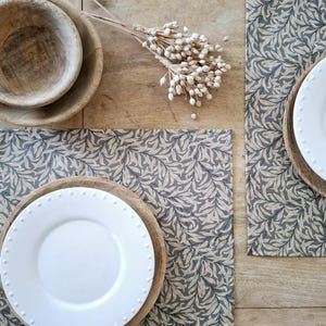 May include: Two placemats with a beige and grey leaf pattern, a white plate on a wooden charger, and a wooden bowl on a wooden table.