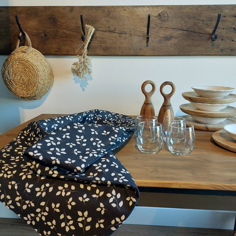 May include: A wooden table displays a dark blue tablecloth with a white leaf pattern, stemless glasses, wooden serving pieces, and stacked white plates. A woven basket and a wooden rack with hooks are visible in the background.