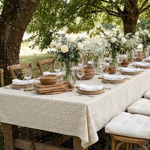 May include: A long wooden table set for a gathering, covered with a floral tablecloth. The table is adorned with white flowers in glass vases, wooden bowls, plates, and wine glasses. Wooden chairs and white cushions complete the outdoor dining scene.