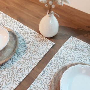 Peut inclure: Deux sets de table blancs avec un motif feuillu brun clair et bleu. Les sets de table sont sur une table en bois avec un vase blanc et des fleurs séchées.