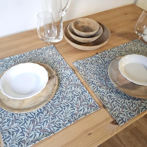 Peut inclure: Mise en place pour le dîner avec deux sets de table à motifs de feuilles bleues et vertes. Chaque set de table comprend une assiette blanche et un dessous de plat en bois. Des bols en bois, une carafe et des verres à vin sont également présents.