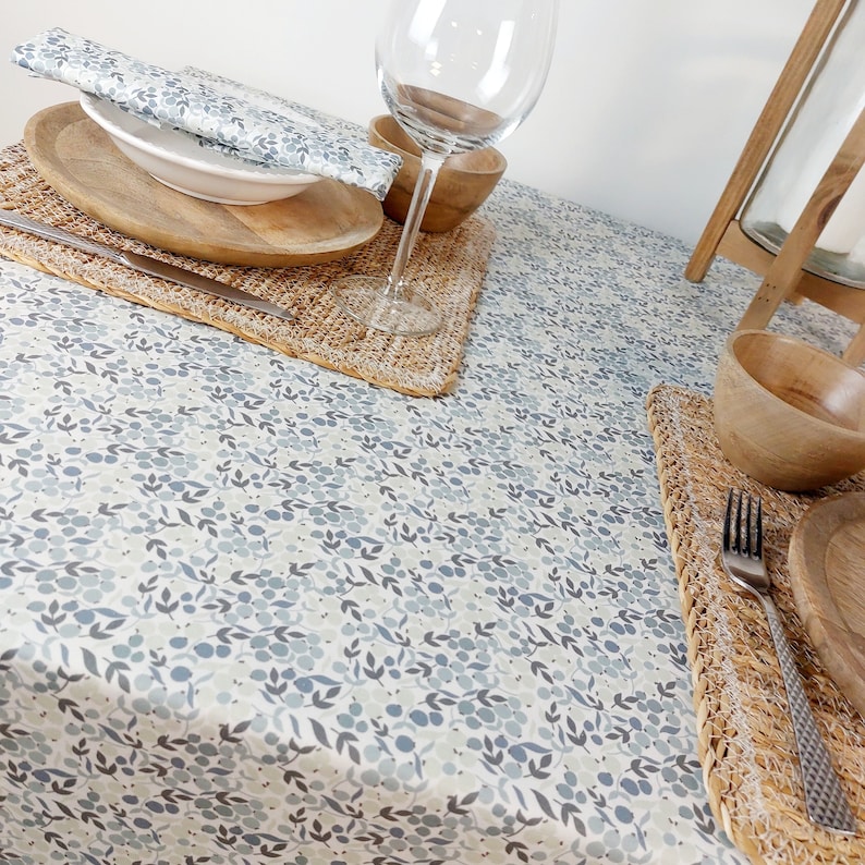 May include: A white tablecloth with a blue and gray floral pattern. The tablecloth is set with a place setting that includes a wooden plate, a bowl, a glass, a fork, and a napkin.