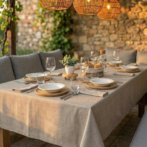 Op de afbeelding: Een eettafel gedekt voor een maaltijd buitenshuis. De tafel is bedekt met een beige linnen tafelkleed en gedekt met witte borden, houten onderzetters, bestek en wijnglazen. Geweven hanglampen hangen erboven.