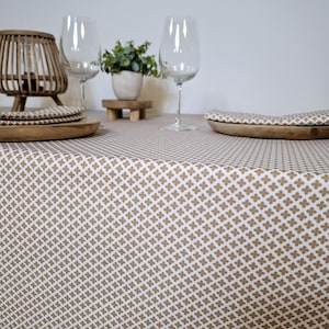 May include: A white tablecloth with a brown geometric pattern. The tablecloth is laid on a table with two wine glasses and a wooden bowl with napkins.