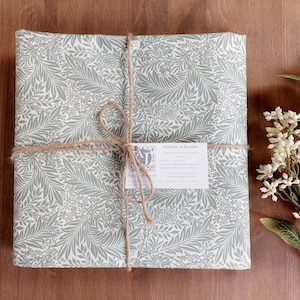 Puede incluir: Un paquete rectangular envuelto en papel con estampado floral verde claro y blanco, atado con hilo de cáñamo. Una pequeña etiqueta blanca con texto negro está sujeta al hilo. El paquete está sobre una superficie de madera.