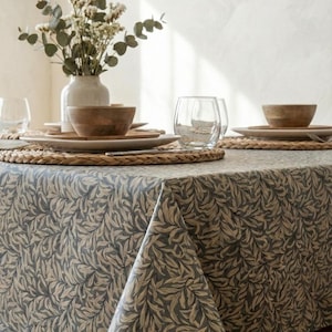 Könnte beinhalten: Ein gedeckter Esstisch mit einer Tischdecke mit blau-beigem Blättermuster. Der Tisch ist mit Holzschalen, Tellern, gewebten Tischsets und klaren Gläsern gedeckt. Eine weiße Vase mit Grünzeug befindet sich im Hintergrund.