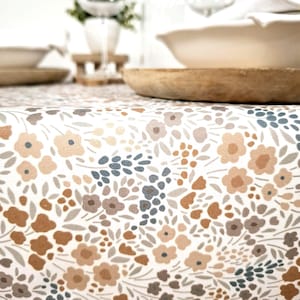 Könnte beinhalten: Nahaufnahme einer floralen Tischdecke mit kleinen Blumen in Braun-, Beige- und Blautönen auf weißem Hintergrund. Die Tischdecke liegt auf einem Tisch mit Holzschalen und einem Glas.