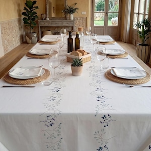 Puede incluir: Una mesa de comedor preparada para una comida, con un mantel blanco con un estampado floral azul. La mesa está puesta con platos, servilletas, vasos y manteles individuales tejidos. También hay botellas de aceite y vinagre, pan y una pequeña planta en maceta.