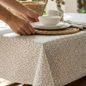 Peut inclure: Une table dressée avec un bol et une assiette blancs sur un dessous de plat en osier. La table est recouverte d'une nappe crème à motifs de feuilles. Un panier en osier et un vase avec des fleurs séchées sont en arrière-plan.