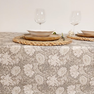 Può includere: Una tovaglia beige con un motivo floreale bianco. Due coperti sono apparecchiati con piatti bianchi, sottopiatti in legno e sottopiatti intrecciati. Due bicchieri di vino trasparenti sono sul tavolo.