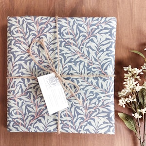 Op de afbeelding: Een cadeau verpakt in blauw en wit bloemenpapier, vastgebonden met touw en een label. Het papier heeft een herhalend patroon van bladeren en takken in lichtblauw en wit, met roze accenten. Het touw is in een strik geknoopt.