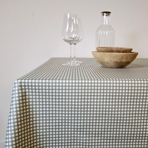 May include: A table set with a green and white gingham tablecloth. Two clear wine glasses, a clear glass bottle with a cork stopper, and two wooden bowls are on the table. The scene is lit with natural light.
