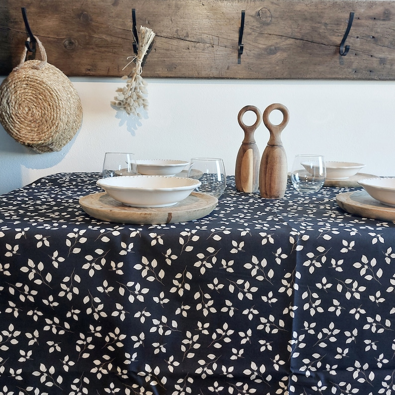 May include: A dining table set with a navy blue tablecloth with a white leaf pattern. The table is set with white bowls, wooden chargers, clear glasses, and wooden salt and pepper grinders. A rustic wooden beam with hooks hangs above.