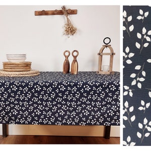 May include: A dark blue tablecloth with a white leaf design. The table is set with white bowls, wooden plates, and a woven placemat. Decorative wooden items and a candle lantern are also on the table.