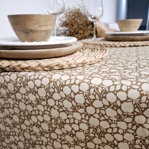 May include: A brown tablecloth with a white floral pattern. The tablecloth is set with a wooden placemat, a white plate, and a wooden bowl.