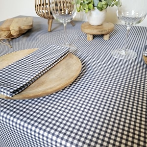 Peut inclure: Une nappe à carreaux noirs et blancs avec une assiette en bois et une serviette pliée dessus. Il y a deux verres à vin et une plante dans un pot blanc sur un support en bois sur la table.