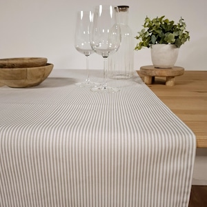 Könnte beinhalten: Ein weißer und grauer gestreifter Tischläufer mit einem dezenten Glanz. Der Läufer liegt auf einem Holztisch mit zwei Weingläsern und einer Glasflasche.