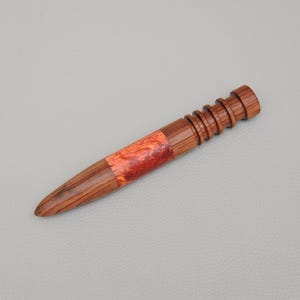 May include: A handcrafted wooden awl with a tapered point and a ridged handle. The awl features a vibrant orange and red resin inlay near the center. The awl is approximately 6 inches long.