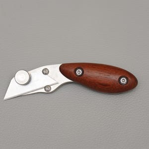 Könnte beinhalten: Ein Cuttermesser mit einer Klinge aus rostfreiem Stahl und einem dunklen Holzgriff. Der Griff ist mit drei silbernen Nieten befestigt. Das Messer hat ein abgerundetes Ende und einen kreisförmigen Klingenverstellmechanismus. Die Klinge ist abgewinkelt.