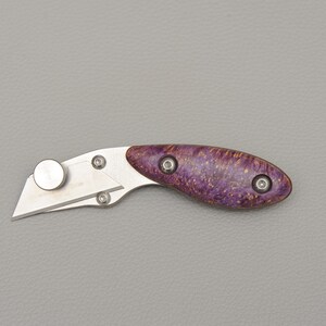 Puede incluir: Un cúter con una cuchilla de acero inoxidable y un mango de madera de burl morado y marrón. El mango está asegurado con tres tornillos plateados. El cuchillo está diseñado para cortar y uso general.