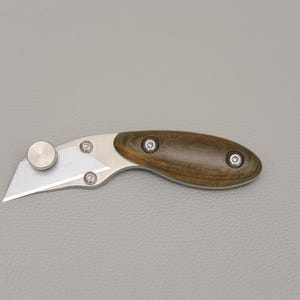 May include: A small utility knife with a silver blade and a wooden handle. The handle is a dark brown color with a wood grain pattern and is secured with three silver rivets. The blade is angled and has a silver thumb stud.