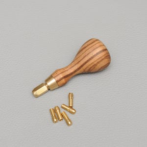 May include: A leathercraft tool with a wooden handle and brass fittings. Several small brass components are positioned nearby. The handle has a striped wood grain pattern. The tool is likely used for leatherwork.