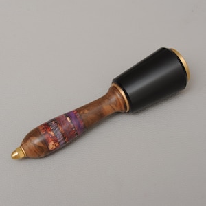 May include: A wooden mallet with a black head and a handle featuring a mix of brown, purple, and red hues. The mallet has a gold-colored band and a gold tip. The tool is designed for woodworking or crafts.