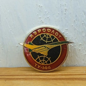 Pode incluir: Um alfinete vermelho, branco e dourado com o texto "АЭРОФЛОТ" e "ТУ-144" em torno de uma imagem preta e verde de um jato supersônico.