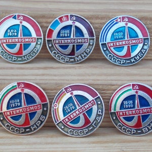 May include: Six round, silver, enamel pins with red, white, blue, and green accents. Each pin features a rocket ship, the text "INTERKOSMOS", and a date. The text "CCCP" is on each pin, followed by a two-letter abbreviation.