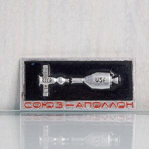 May include: A silver metal pin with a black background depicting a Soyuz spacecraft docked with an Apollo spacecraft. The text "СОЮЗ-АПОЛЛОН" is written in red letters below the image.