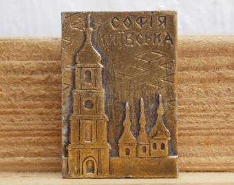 Puede incluir: Un souvenir de metal dorado que representa la Catedral de Santa Sofía en Kiev, Ucrania. El texto "СОФІЯ КИЇВСЬКА" está grabado en la parte superior del souvenir.