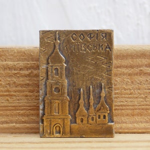 Może przedstawiać: Złoty metalowy pamiątkowy znaczek z wizerunkiem Soboru św. Zofii w Kijowie na Ukrainie. Na górze pamiątki wygrawerowano tekst "СОФІЯ КИЇВСЬКА".