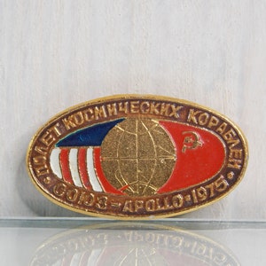 May include: A gold-colored oval pin with the text "ПОЛЕТ КОСМИЧЕСКИХ КОРАБЛЕЙ СССР - АПОЛЛО - 1975" in Russian. The pin features a globe with a red and blue flag on each side.