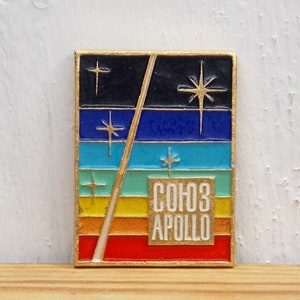 May include: A gold-colored pin with a rainbow design and a white rectangle with the text "СОЮЗ АПОЛЛО" in black letters. The pin features a diagonal line and stars.