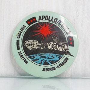 May include: A round, blue and white pin with a black and white image of two spacecraft docked together. The pin has the text "APOLLO/SOYUZ" and "STAFFORD - BRAND - SLAYTON" on the top half and "LEONOV - KUBASOV" on the bottom half.