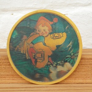 May include: A round, yellow-rimmed coaster with a colorful image of a cartoon character wearing a red hat and holding a yellow object. The background is a green and blue landscape.