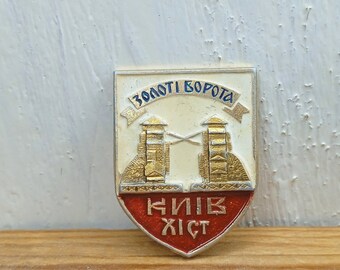 Puede incluir: Un pin de esmalte dorado y blanco con forma de escudo. El escudo presenta una representación de la Puerta Dorada, un hito histórico en Kiev, Ucrania. El texto "Золоті ворота" está escrito en azul sobre la puerta, y "КИЇВ ХІСТ" está escrito en rojo debajo de la puerta.